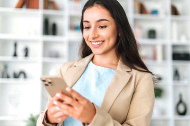 Kadın akıllı telefon kullanıyor. Takım elbiseli başarılı Arap ya da Hintli kadın, ofiste duruyor, cep telefonu tutuyor, sosyal medyada arkadaşlarıyla veya müşterileriyle mesajlaşıyor, çevrimiçi sipariş veriyor, gülümsüyor.