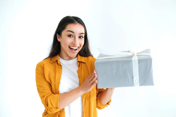 Gift box. Surprised pretty brunette indian or arabian woman in casual orange shirt, stands on isolated white background, holding big gift box, excited looking at the camera and smiles friendly