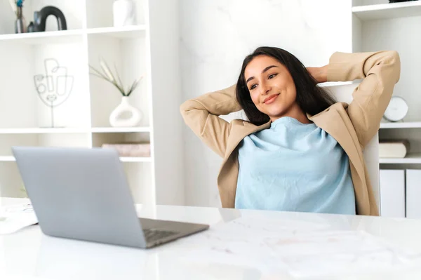 Satisfied carefree woman of indian or arabian nationality, sitting relaxed in a chair at her desk with her hands behind her head, looking at her laptop, taking a break from work tasks, smiling