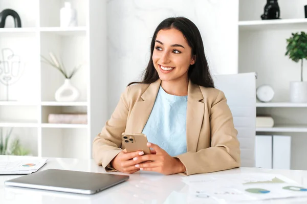 Attractive arabian or indian business woman, dressed in elegant clothes, company employee, sit at workplace in the office, using her smartphone, messaging with friends or clients, looks away, smiling