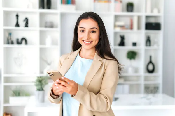 Attractive successful arabian or indian business woman in a suit, stands in the office, holding mobile phone, texting in social media with friends or clients, making online order,looks at camera,smile