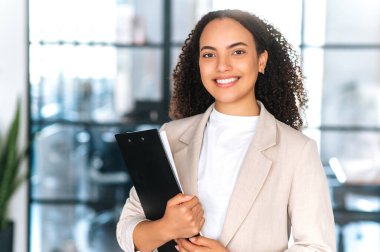 Pozitif, kıvırcık saçlı, zarif giysiler içinde İspanyol bir iş kadınının portresi. Sekreter, iş kadını, modern ofiste elinde belgeler tutarken kameraya gülümse.