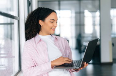 Cihazları ve aletleri kullanarak. Güzel, başarılı Brezilyalı ya da Latin kıvırcık saçlı kadın, yazılım yöneticisi, şirket çalışanı, modern ofiste elinde bir dizüstü bilgisayarla duruyor, ona bakıyor, gülümsüyor.