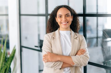 Güzel, başarılı, kıvırcık saçlı, zarif elbiseli bir iş kadını, şirket müdürü, şirketin CEO 'su, kollarını kavuşturmuş, kameraya bakıyor, gülümsüyor.