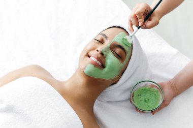 Kadın yüzüne kilden bir maske takma prosedürü. Güzel bir Brezilyalı ya da İspanyol genç kadının yüzünün yarısına yeşil yüz maskesi takan terapistin yakın çekimi. Cilt bakımı kavramı