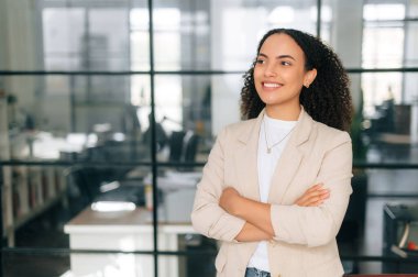 Başarılı, kendine güvenen, kıvırcık saçlı bir iş kadınının resmi giysiler içinde modern yaratıcı bir ofiste kollarını kavuşturmuş gülümseyen bir şirket müdürünün fotoğrafı.