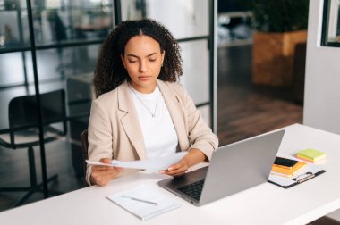 Ciddi kendine güvenen Brezilyalı ya da İspanyol kıvırcık saçlı iş kadını, şirket CEO 'su, finans müdürü, modern ofiste bir çalışma masasında oturuyor, bir dizüstü bilgisayarda çalışıyor, finansal belgeleri inceliyor.