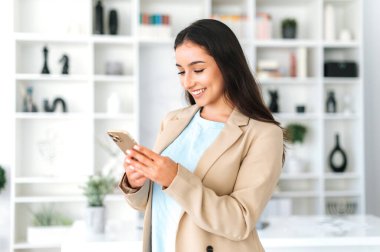 Cihazları kullanarak. Takım elbiseli mutlu Arap ya da Hintli kadın, ofiste duruyor, cep telefonu tutuyor, sosyal medyada arkadaşlarıyla veya müşterileriyle mesajlaşıyor, online sipariş veriyor, gülümsüyor.