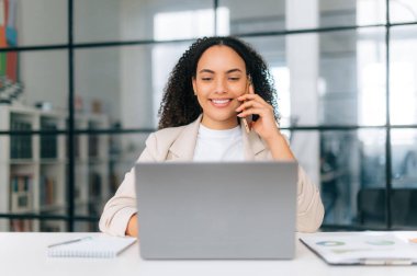 Meşgul, muhteşem bir Brezilyalı ya da İspanyol iş kadını, CEO, modern ofiste oturuyor, bir dizüstü bilgisayarda çalışıyor, müşteriyle ya da meslektaşıyla akıllı telefondan konuşuyor, arkadaşça gülümsüyor.