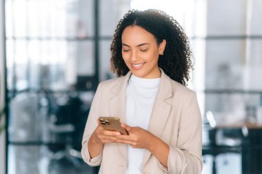 Cihazları kullanarak. Muhteşem başarılı Brezilyalı ya da İspanyol zarif kadın, şirket yöneticisi, sekreteri, modern ofis alanında duruyor, akıllı telefonu müşteri ya da partnerle sohbet etmek için kullanıyor, daktilo ediyor, gülümsüyor.