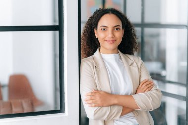 Kendine güvenen, başarılı İspanyol ya da kıvırcık saçlı Brezilyalı stil bir kadının portresi modern bir ofiste kollarını kavuşturup kameraya bakıyor ve dostça gülümsüyor.