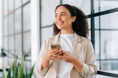 İnternette sohbet ediyoruz. Kıvırcık saçlı pozitif çok ırklı genç bir kadın, modern bir ofiste duruyor, internet üzerinden arkadaşlarıyla veya müşterileriyle mesajlaşıyor, haberleri kontrol ediyor, yüzünü çeviriyor, gülümsüyor.