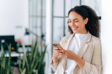 Başarılı, pozitif Brezilyalı ya da zarif ceketli kıvırcık saçlı İspanyol genç kadın, modern bir ofiste duruyor, arkadaşlarıyla ya da müşterileriyle mesajlaşıyor, internette haberlere bakıyor, gülümsüyor.