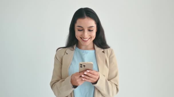 用小玩意积极漂亮的印度或阿拉伯女人 穿着雅致的衣服 用智能手机与朋友聊天 浏览互联网 站在孤立的白色背景上 快乐地微笑 — 图库视频影像