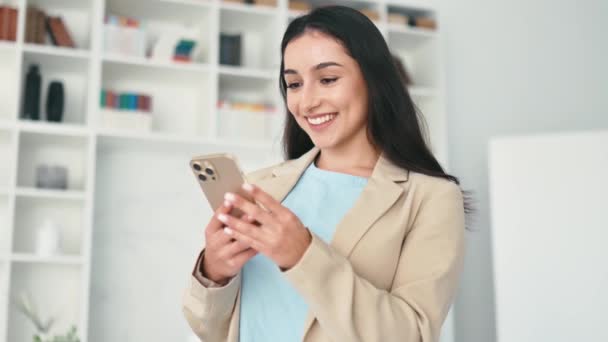 用小玩意穿着西装的快乐的成功的阿拉伯或印度女人 站在办公室里 拿着手机 与朋友或客户在社交媒体上发短信 在网上下订单 — 图库视频影像