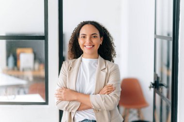 Çekici, kendine güvenen, başarılı, İspanyol asıllı, kıvırcık saçlı, modern bir ofiste kollarını kavuşturmuş kameraya bakıp gülümseyen genç bir kadın.