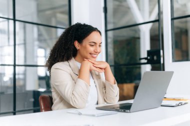 Güzel Brezilyalı ya da İspanyol kıvırcık saçlı, zarif elbiseli iş kadını, CEO, İK müdürü, modern bir ofiste dizüstü bilgisayarla oturuyor, cihaz ekranına bakıyor, arkadaşça gülümsüyor.