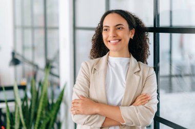 Çekici başarılı Latin ya da Brezilyalı kıvırcık iş kadını, zarif giysiler içinde, şirket pazarlama müdürü, şirketin CEO 'su, modern ofiste kollarını kavuşturmuş, yüzünü çevirmiş, gülümsüyor.