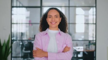 Ofis çalışanı. Kendine güveni tam, başarılı İspanyol ya da Brezilyalı stil kıvırcık saçlı genç bir kadın modern bir ofiste kollarını kavuşturup kameraya bakıyor ve gülümsüyor.