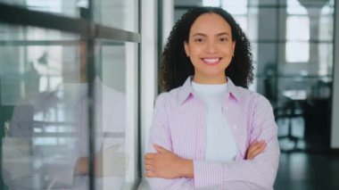 Kendine güvenen, başarılı, İspanyol asıllı, stil sahibi, kıvırcık saçlı, modern bir ofiste kolları çapraz duran, kameraya bakan, arkadaşça gülümseyen bir kadının kadın portresi.