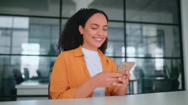 驚くべき幸せなヒスパニックまたはブラジルのカーリーハイレッド女性オレンジ色のシャツで オフィスの職場に座って 彼女の携帯電話を使用して 友人とのメッセージング ソーシャルメディアの閲覧 カメラで笑顔 ストック動画