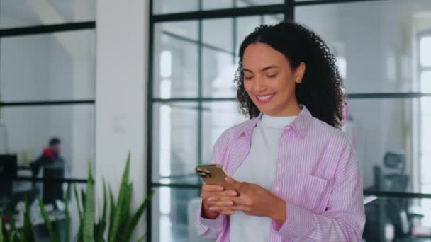 ポジティブなブラジル人またはラテン系のカーリービジネス女性 コーポレートマネージャー デザイナーは ガジェットを使用してスマートフォンで近代的なオフィスを歩いて それを見て テキストを入力し ストック映像
