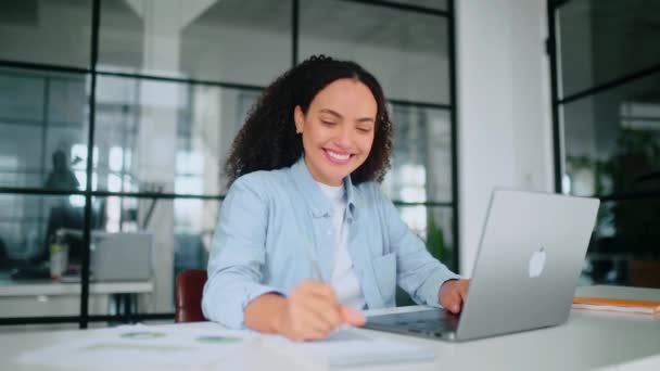 一个穿着淡蓝色衬衫 精力充沛的漂亮女人 一个公司的员工 坐在一个现代化的办公室的办公桌前 在笔记本电脑上工作 在笔记本上记笔记 微笑着 视频剪辑