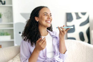 Neşeli, güzel Brezilyalı ya da lavanta rengi gömlekli İspanyol kıvırcık bir kadın, elinde akıllı bir telefonla oturuyor, hoparlörden konuşuyor ya da sesli mesaj kaydediyor, gülümsüyor.