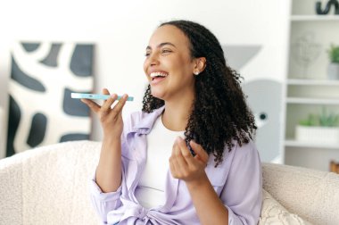 Muhteşem, pozitif Brezilyalı ya da İspanyol kıvırcık, lavanta gömlekli, elinde akıllı bir telefonla oturan, hoparlörden konuşan ya da sesli mesaj kaydeden, gülümseyen bir kadın.