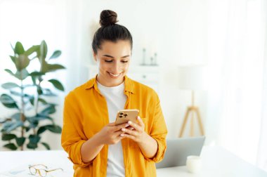 Ofiste ya da evde dururken cep telefonu tutan turuncu tişörtlü mutlu Arap ya da Hintli genç kadın, sosyal medyada mesajlaşıyor, internette geziniyor, uygulamalarla ürünler sipariş ediyor.