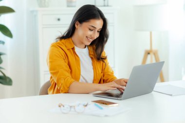 Pozitif başarılı esmer Arap ya da Hint kökenli genç kadın, bilişim uzmanı, ürün yöneticisi bir çalışma masasında oturuyor, bilgisayarında çalışıyor, cihaz ekranına bakıyor, mutlu bir şekilde gülümsüyor.