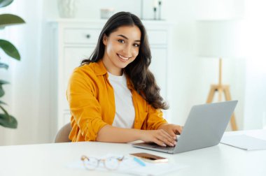 Turuncu gömlekli güzel, başarılı esmer Arap ya da Hintli genç kadın, bilişim uzmanı, ürün yöneticisi bir iş yerinde dizüstü bilgisayarda çalışıyor, kameraya bakıyor, arkadaşça gülümsüyor.