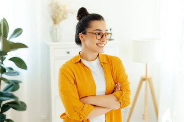 Tarz sahibi, muhteşem Arap ya da Hint kökenli, gözlüklü, şirket çalışanı, yaratıcı yönetici kollarını kavuşturup gülümseyerek çalışma masasının yanında duran turuncu gömlekli genç bir kadın.