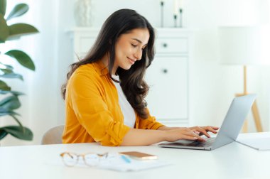 Odaklanmış güzel esmer Arap ya da Hintli genç kadın, bilişim uzmanı, ürün yöneticisi, web tasarımcısı, bir dizüstü bilgisayarda çalışıyor, dizüstü bilgisayara bakıyor, gülümsüyor.