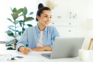 Olumlu, kendine güvenen Arap ya da Hintli iş kadını, proje yöneticisi, şirket CEO 'su, bir dizüstü bilgisayarda çalışıyor, planlama stratejisi ve iş planı yapıyor, notlar alıyor, uzaklara bakıyor ve gülümsüyor.