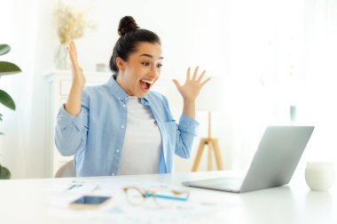 Şaşırtıcı mutlu Hintli ya da Arap başarılı iş kadını, bir iş yerinde oturuyor, bir dizüstü bilgisayara bakıyor, başarıyla seviniyor, iyi iş sonuçları, gülümsüyor, elleriyle hareket ediyor