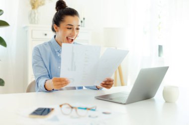 Mutlu, heyecanlı Hintli ya da Arap başarılı iş kadını bir iş yerinde oturuyor, finansal belgeleri analiz ediyor, başarıya seviniyor, iyi iş çıkartıyor, gülümsüyor.