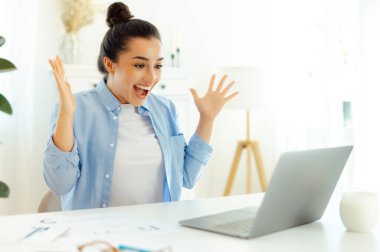 Beklenmedik haberler. Şaşırmış mutlu Hintli ya da Arap başarılı iş kadını, bir iş yerinde oturuyor, şaşkınlıkla dizüstü bilgisayara bakıyor, başarıyla seviniyor, gülümsüyor, elleriyle hareket ediyor.