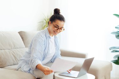 Gözlüklü, serbest çalışan, dijital çalışan, web tasarımcısı, oturma odasında kanepede otururken dizüstü bilgisayar kullanan, gülümseyen mutlu Hintli ya da Arap kadın. Çevrimiçi eğitim ve çalışma