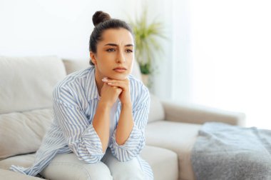 Gündelik giysiler içinde üzgün Arap ya da Hintli genç bir kadın, oturma odasında bir kanepede oturuyor, üzgün, yorgun ve yalnız hissediyor, depresyon geçiriyor, kişisel sorunları ya da boşanması için endişeleniyor, güceniyor.