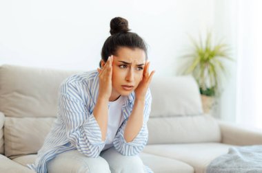 Zor bir iş gününden sonra oturma odasındaki koltukta oturan, baş ağrısı, baş dönmesi, baş dönmesi, migren, tedavi ve dinlenmeye ihtiyacı olan, masaj yaptıran esmer kadın. Stres, baş ağrısı, kramp.