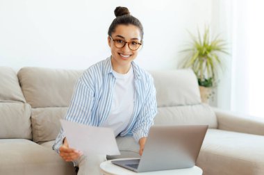 Hindistan ya da Arap kökenli gözlüklü mutlu, bağımsız bir kadın oturma odasında bir kanepede oturmuş dizüstü bilgisayar kullanıyor, elinde belgeler, kameraya bakıyor, gülümsüyor.