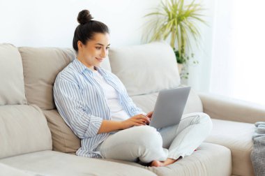 Online iş. Mutlu Hintli ya da Arap esmer kadın, rahat bir kanepede oturuyor, oturma odasında, dizüstü bilgisayarını uzak bir proje için kullanıyor, gülümsüyor.