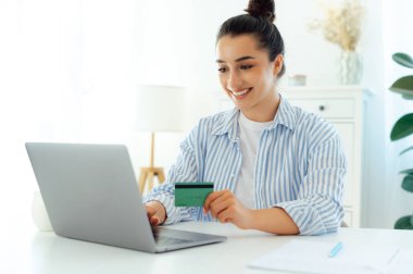 Güzel, mutlu Hintli ya da Arap genç bir kadın elinde bir kredi kartı tutarken ve bir dizüstü bilgisayarla masada otururken internetten alışveriş yaparken gülümserken. Para transferi, e-bankacılık, online alışveriş