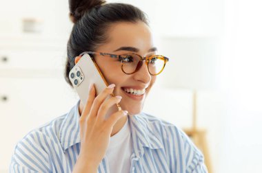 Mavi tişörtlü başarılı beyaz iş kadınının yakın plan fotoğrafı. Ofisinde çalışırken bir müşteriyle ya da ortağıyla telefonda konuşuyor. Başka tarafa bakıyor, gülümsüyor.