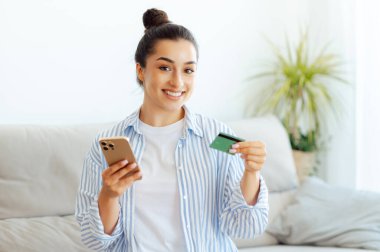 E-bankacılık konsepti, online para harcamak. Mutlu genç bir kadın, akıllı telefon ve kredi kartını internet uygulamasında online ödeme için kullanıyor, online alışveriş yapıyor, para transfer ediyor, gülümsüyor.