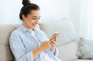 Sosyal medya, aletler. Rahat bir koltukta oturup arkadaşlarıyla online iletişim kurmak için akıllı bir telefonda yazan, komik videolar izleyen, gülümseyen mutlu, beyaz bir kadın.