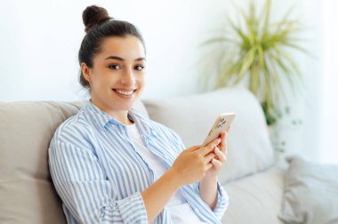 Neşeli, mutlu, güzel Hintli ya da Arap kadın gündelik kıyafetlerle rahat koltukta oturuyor, akıllı telefon mesajları kullanıyor arkadaşlarıyla internette geziniyor, çevrimiçi dükkanlara bakıyor ve kameraya gülüyor.