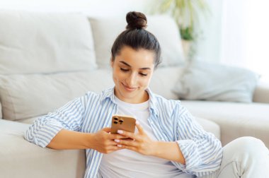 Pozitif Modern Kafkas esmer kadın, günlük giysiler içinde, oturma odasında bir kanepenin yanında oturuyor, akıllı telefon mesajları kullanıyor arkadaşlarıyla internette geziniyor, online mağazalara bakıyor, gülümsüyor.