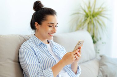 Neşeli, mutlu, Hintli ya da Arap elbiseli güzel bir kadın, rahat bir koltukta oturup arkadaşlarıyla mesajlaşıyor, komik videolar izliyor, online mağazalara bakıyor, gülümsüyor.
